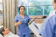 Student Nurse in blue scrubs holding a clipboard, speaking to colleagues in a hospital room with a Nursing Mannequin lying in bed.